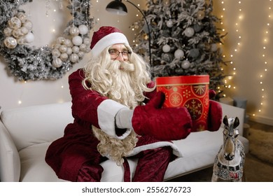 Santa claus holding red christmas gift box sitting on sofa with rocking horse near decorated christmas tree - Powered by Shutterstock