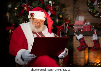 Santa Claus with laptop. Fireplace and Christmas tree. Festive interior inside wooden house, New Year's cheerful mood Spirit of Christmas. Senior man with real white beard cosplay Father Christmas. - Powered by Shutterstock