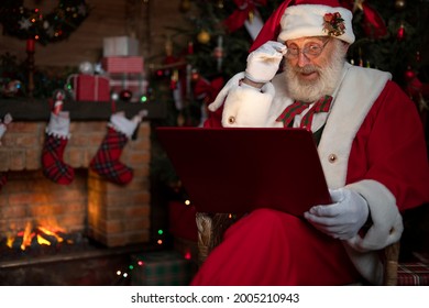 Santa Claus with laptop. Fireplace and Christmas tree. Festive interior inside wooden house, New Year's cheerful mood Spirit of Christmas. Senior man with real white beard cosplay Father Christmas. - Powered by Shutterstock