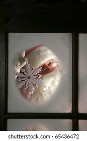 Santa Claus Looks Out Through The Snow And Fog On His Workshop Window In The North Pole To See Outside Check The Weather While He Holds Up A Large Snow Flake  On December 24th, Christmas Eve