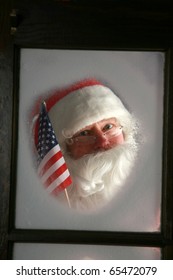 Santa Claus Looks Out Through The Snow And Fog On His Workshop Window In The North Pole To See Outside Check The Weather While He Holds Up An American Flag On December 24th, Christmas Eve