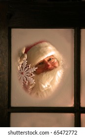 Santa Claus Looks Out Through The Snow And Fog On His Workshop Window In The North Pole To See Outside Check The Weather While He Holds Up A Large Snow Flake On December 24th, Christmas Eve