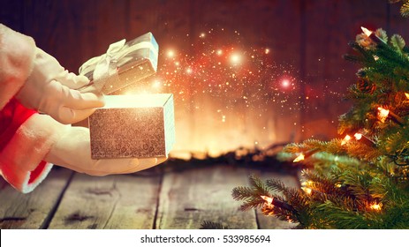 Santa Claus opens gift box, holding a gift in his hands over wooden background with blinking garland and Christmas tree. Xmas scene with magic gift, wishes.
