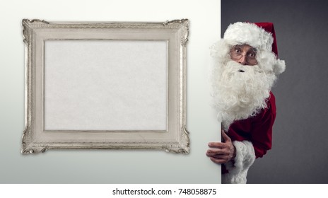 Santa Claus Peeking Behind A Wall And Blank Decorative Frame, Christmas And Holidays Concept