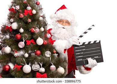 Santa Claus posing behind a Christmas tree and holding a movie clapperboard isolated on white background
