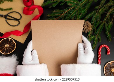 Santa Claus Reading Letters from Children on Desk. Top View of Jolly Father Hands Holding a Blank Brown Paper Page. View from Above of Wood Table with Ornament, Christmas Decoration. Mock Up - Powered by Shutterstock