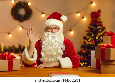 Santa Claus in red suit and hat with white beard sitting at table with presents waving hand, saying hello during video call or greeting subscribers on live stream, vlog or channel - Powered by Shutterstock