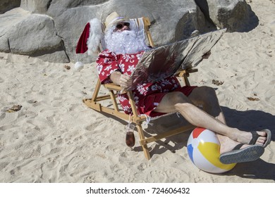 Santa Claus In Red Swimming Trunks Ans Hawaiian Shirt Lounging On Sandy Beach With Straw Hat And Sunglasses Using Reflector To Tan.