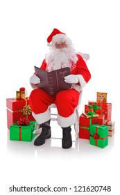 Santa Claus Sat In A Rocking Chair Surrounded By Gift Wrapped Presents As He Reads The Naught Or Nice List, Isolated On A White Background.