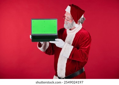 Santa Claus showcasing laptop with green screen ideal for adding custom content. Elderly man standing against red background wearing Santa costume holding laptop - Powered by Shutterstock