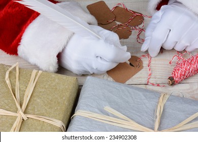 Santa Claus Sitting At His Desk Writing Gift Tags For Christmas Presents. Horizontal Closeup Showing Gifts And Santa's Hands Only.