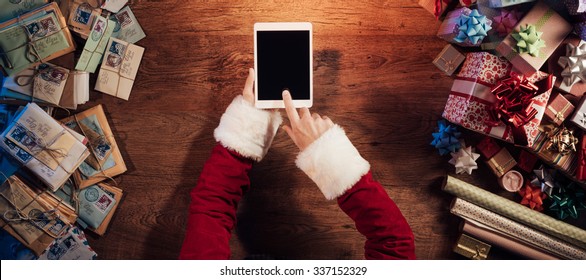 Santa Claus sitting at his desk and using a digital touch screen tablet surrounded by colorful Christmas gifts - Powered by Shutterstock