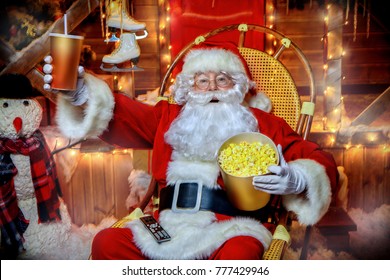 Santa Claus sitting on his armchair eating popcorn, drinking soda and watching a Christmas movie. Entertainment and cinema concept. Merry Christmas and Happy New Year.