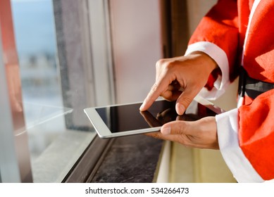 Santa Claus Standing At Window And Using Apps Resize On A Touch Screen Tablet, Christmas And Technology Concept