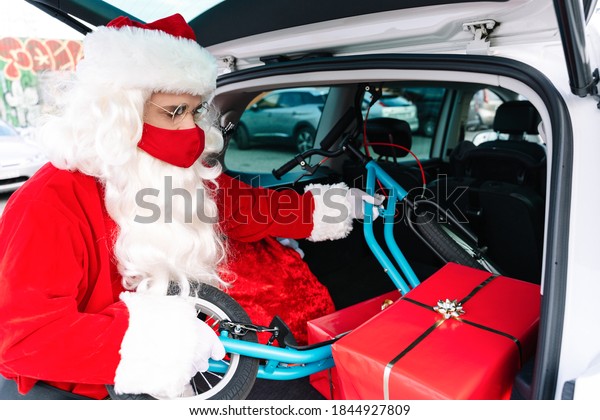 Santa Claus with the trunk of the car open full of\
gifts taking out a children\'s scooter with big wheels to deliver\
it, he is wearing a face mask due to the coronavirus covid19 in\
Christmas 2020