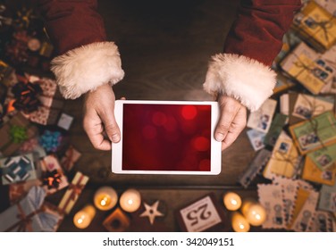 Santa Claus using a digital touch screen tablet, hands close up, top view, desktop with letters and Christmas gifts on background