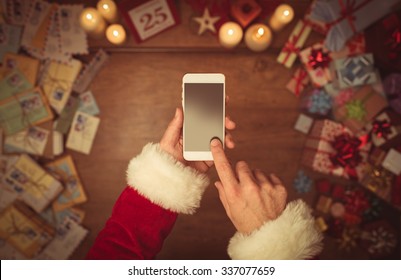 Santa Claus using a touch screen smart phone, hands close up, top view, desktop with gifts and Christmas letters on background - Powered by Shutterstock