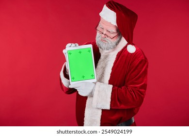 Santa Claus wearing traditional costume, holding a tablet with green screen background, standing against vibrant red background. Perfect for holiday and Christmas-themed projects - Powered by Shutterstock