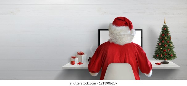 Santa Claus working on a computer in his office during Christmas holidays. Empty space on wall for text.