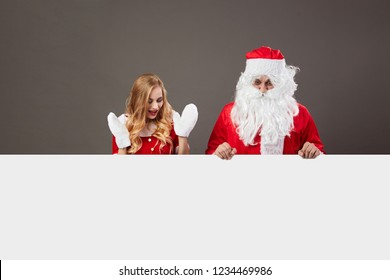 Santa Claus And Young Beautiful Mrs. Claus Stand Behind A White Canvas And Look Surprised On The Gray Background.