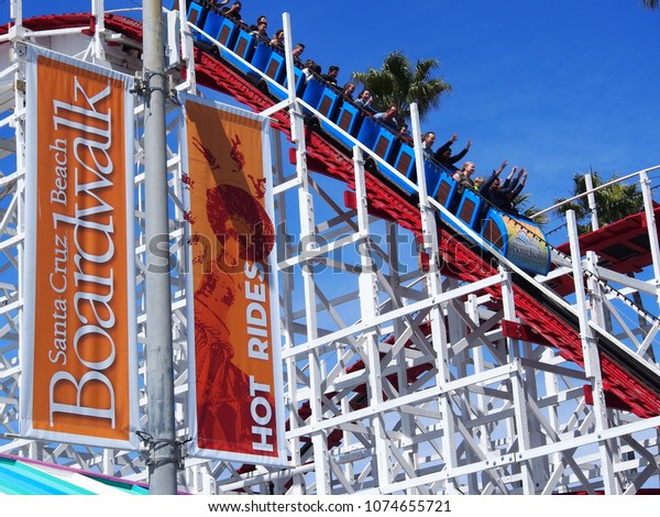 Santa Cruz, California / USA - Circa April 2018:\
Santa Cruz Boardwalk Banner Sign with Guests Riding the Famous\
Giant Dipper Red and White Wooden Roller Coaster in Background,\
Hands Raised in the Air
