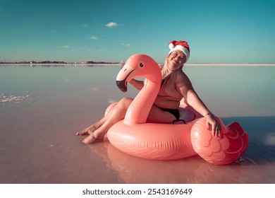 Santa Flamingo Christmas woman Riding Inflatable Flamingo Pink Lake. A woman wearing a Santa hat sits on a pink flamingo float in a body of water. He is smiling.