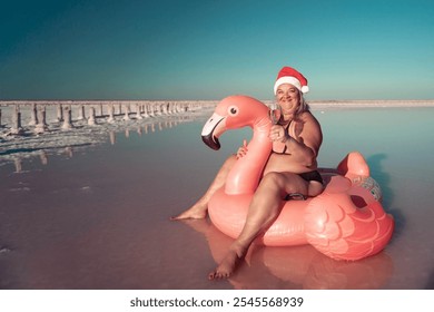 Santa Flamingo Christmas woman Riding Inflatable Flamingo Pink Lake. A woman wearing a Santa hat sits on a pink flamingo float in a body of water. He is smiling.