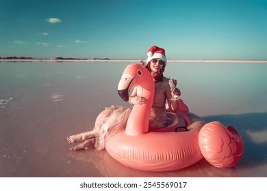 Santa Flamingo Christmas woman Riding Inflatable Flamingo Pink Lake. A woman wearing a Santa hat sits on a pink flamingo float in a body of water. He is smiling.