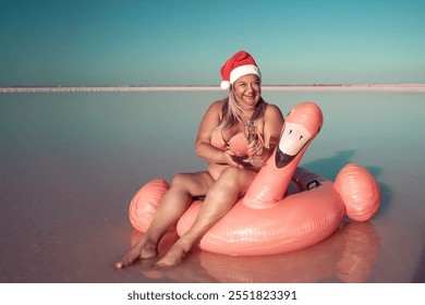 Santa Flamingo Christmas woman Riding Inflatable Flamingo Pink Lake. A woman wearing a Santa hat sits on a pink flamingo float in a body of water. He is smiling.