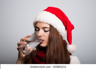 Santa Girl Drinking A Glass Of Water. Christmas Hat Isolated Portrait Of A Woman On A Gray Background.