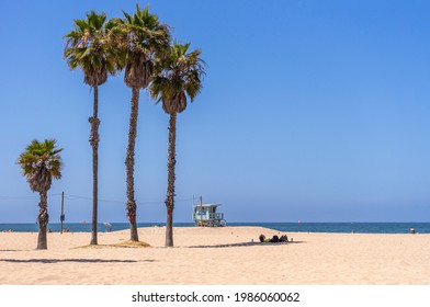 Santa Monica, CA, USA - June 20, 2013: Couple Rests On Sandy Beach In Palm Tree Shadow Under Blue Sky. Lifeguards Lookout Shack In Back At Dark Blue Water.