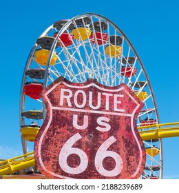 Santa Monica, California, USA - 14 May 2022: A Large Dominant Weathered Red Sign Marks The Start Of Historic Route 66, The Mother Road. A Colourful Fairground Ferris Wheel With Clear Blue Copy Space.