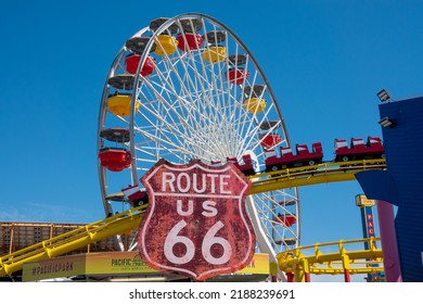 Santa Monica, California, USA - 14 May 2022: A Large Dominant Weathered Red Sign Marks The Start Of Historic Route 66, The Mother Road. A Colourful Fairground Ferris Wheel With Clear Blue Copy Space.