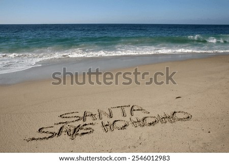 Santa says Ho Ho Ho. Santa Laughs while writing words in wet sand on a beach. The words Santa Enjoys a Beach Vacation by the ocean in the sand. Christmas Vacation. Santa Claus Enjoys the West Coast.