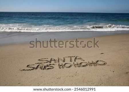 Santa says Ho Ho Ho. Santa Laughs while writing words in wet sand on a beach. The words Santa Enjoys a Beach Vacation by the ocean in the sand. Christmas Vacation. Santa Claus Enjoys the West Coast.