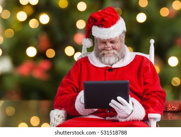 Santa using digital tablet while sitting on a chair against bokeh lights