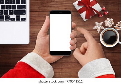 Santa's Hand Holding And Touching Black Phone With Empty Screen At Wooden Desk Surrounded With Laptop, Gift Box, Coffee And Cakes