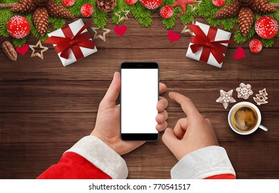 Santa's Hand Holding And Touching Black Phone With Empty Screen At Wooden Desk With Christmas Decoration, Gift Boxes, Coffee And Cakes. Top View