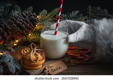 Santa's hand reaches for a glass of milk with cookies, Christmas tree, pine cones, heart-shaped lollipops and a garland on a wooden table, close-up side view. Christmas tradition concept. - Powered by Shutterstock