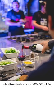 Santiago, Chile - Circa 2017: Hand Serving A Drinking Glass Of Red Wine With A Bottle In A Large Metal Table In A Kitchen With More People And Blue Light