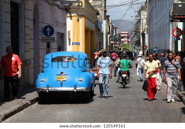 SANTIAGO, CUBA - FEBRUARY 10: People walk past old\
car on February 10, 2011 in Santiago, Cuba. Recent law change\
allows the Cubans to trade cars again. Cars in Cuba are old because\
of the old law.