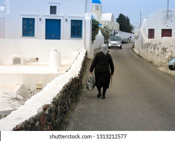 Santorini Island, Greece - April 12, 2018: A Typical Old Lady Widow Walks Through The Alley Carrying Your Shopping Bag. View Of The Street Of The Village, Surrounded By The Old White Houses.  