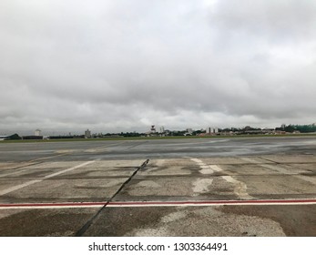 Sao Paulo, Brazil - February 25, 2019: Congonhas Airport, Rainy Day, Sky Covered By Clouds, Wet Lane, A White Plane Landing On The Concrete Floor. Architecture From The Airport To The Background.