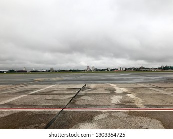 Sao Paulo, Brazil - February 25, 2019: Congonhas Airport, Rainy Day, Sky Covered By Clouds, Wet Lane, A White Plane Landing On The Concrete Floor. Architecture From The Airport To The Background.