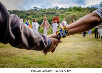 Sao Paulo, SP, Brazil - June 26 2022: Caucasian Male Hands With Indigenous Bracelet Together, In The Background Indigenous People With And Without Headdress Holding Hands In A Circle.