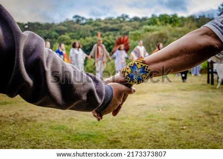 Sao Paulo, SP, Brazil - June 26 2022: Caucasian male hands with indigenous bracelet together, in the background indigenous people with and without headdress holding hands in a circle.