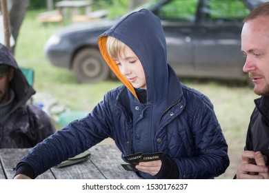 Saratov, Russia - 07/06/2017: A Portrait Of A Small Boy Playing Cards Outside In The Forest With His Family And Friends.