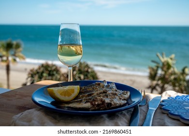 Sardines espeto prepared on skewers and open flame on fireplace with olive trees wood, served outdoor with lemon and glass of fino sherry wine and view on blue sea, Malaga, Andalusia, summer vacation 