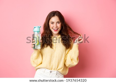 Sassy attractive woman looking confident at camera, holding personal bottle with lemon water healthy drink, standing against pink background