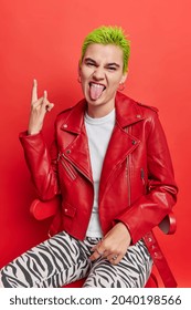 Sassy punk girl sticks out tongue shows heavy metal sign gesture feels cool has trendy green hairstyle wears leather jacket sits on chair isolated over red background enjoys rock music. Youth concept - Shutterstock ID 2040198566